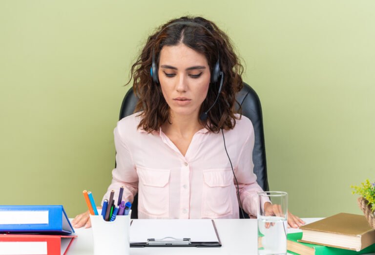 confident-pretty-caucasian-female-call-center-operator-headphones-sitting-desk-with-office-tools-looking-clipboard