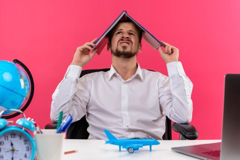 handsome-businessman-white-shirt-with-globe-holding-notebook-his-head-looking-confused-sitting-table-offise-pink-background (1)